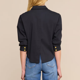 A woman shown from the back wearing a black long-sleeve button-up shirt with gold button accents at the cuffs, paired with blue jeans, against a solid beige background.