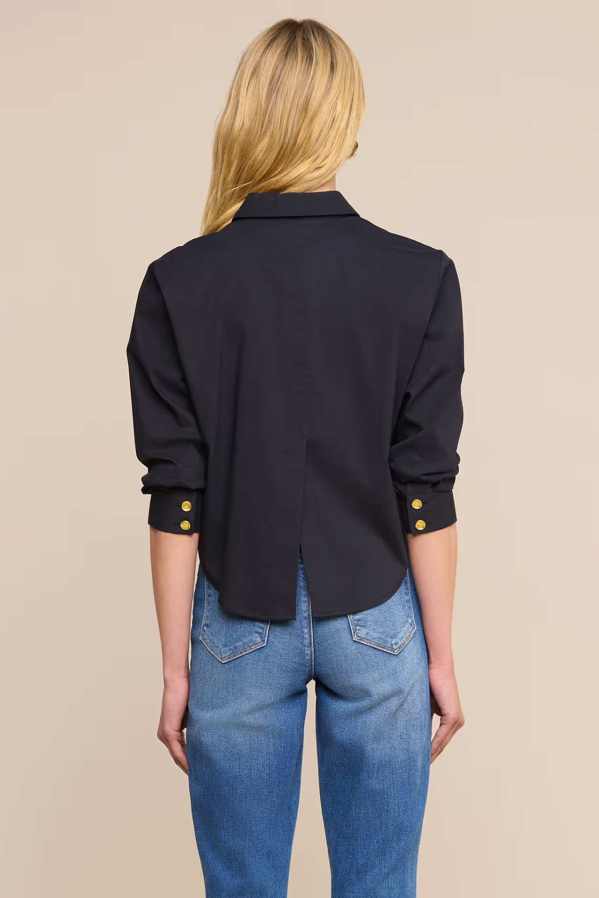 A woman shown from the back wearing a black long-sleeve button-up shirt with gold button accents at the cuffs, paired with blue jeans, against a solid beige background.