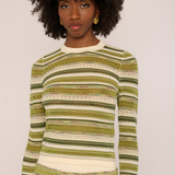 A woman with a dark curly afro hairstyle wearing a long-sleeve knit sweater with horizontal stripes in olive green, beige, and white, set against a plain light background.