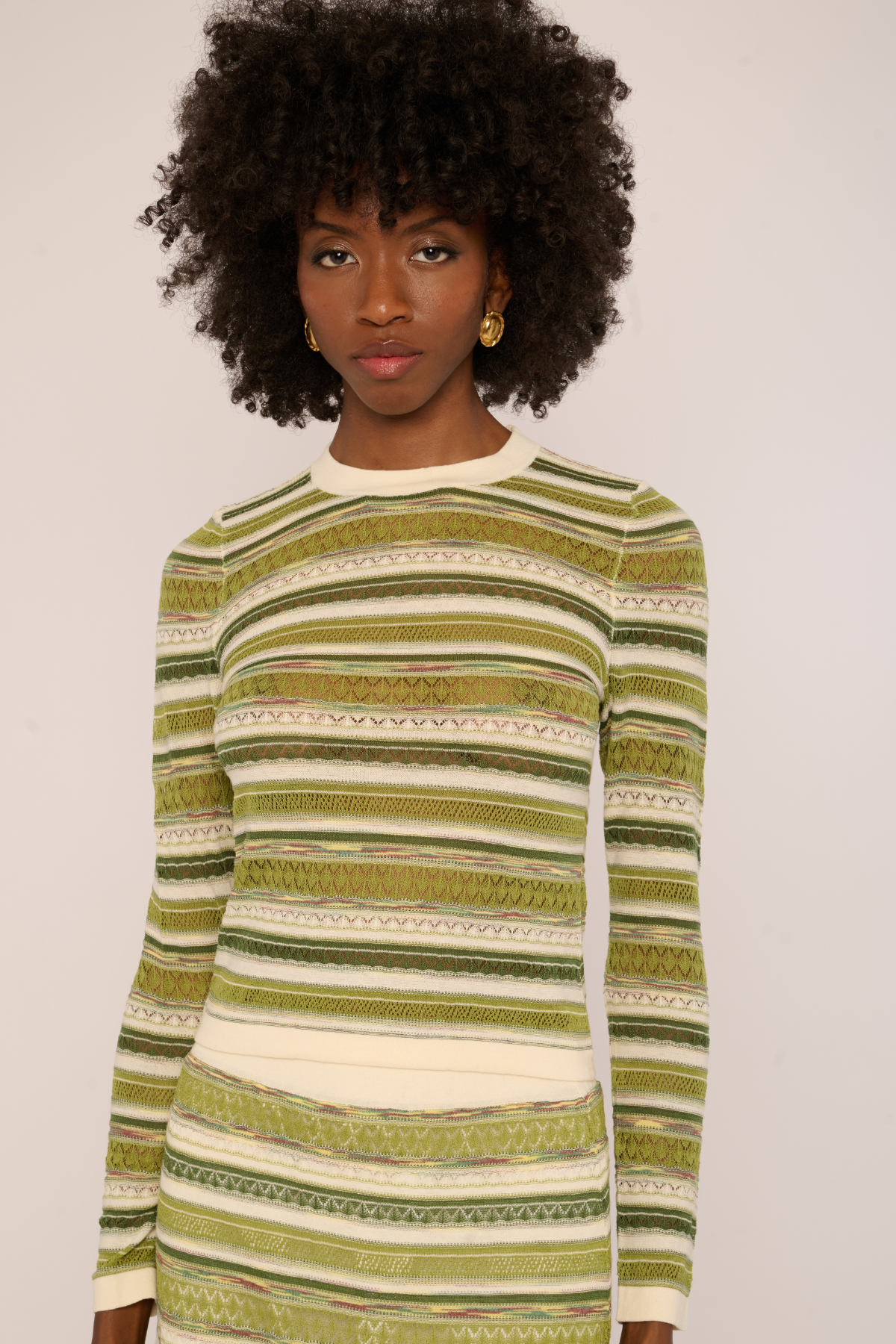 A woman with a dark curly afro hairstyle wearing a long-sleeve knit sweater with horizontal stripes in olive green, beige, and white, set against a plain light background.