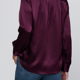 A woman wearing a deep purple satin blouse with a contrasting velvet collar and shoulders, paired with blue jeans, shown from the back against a light gray background.