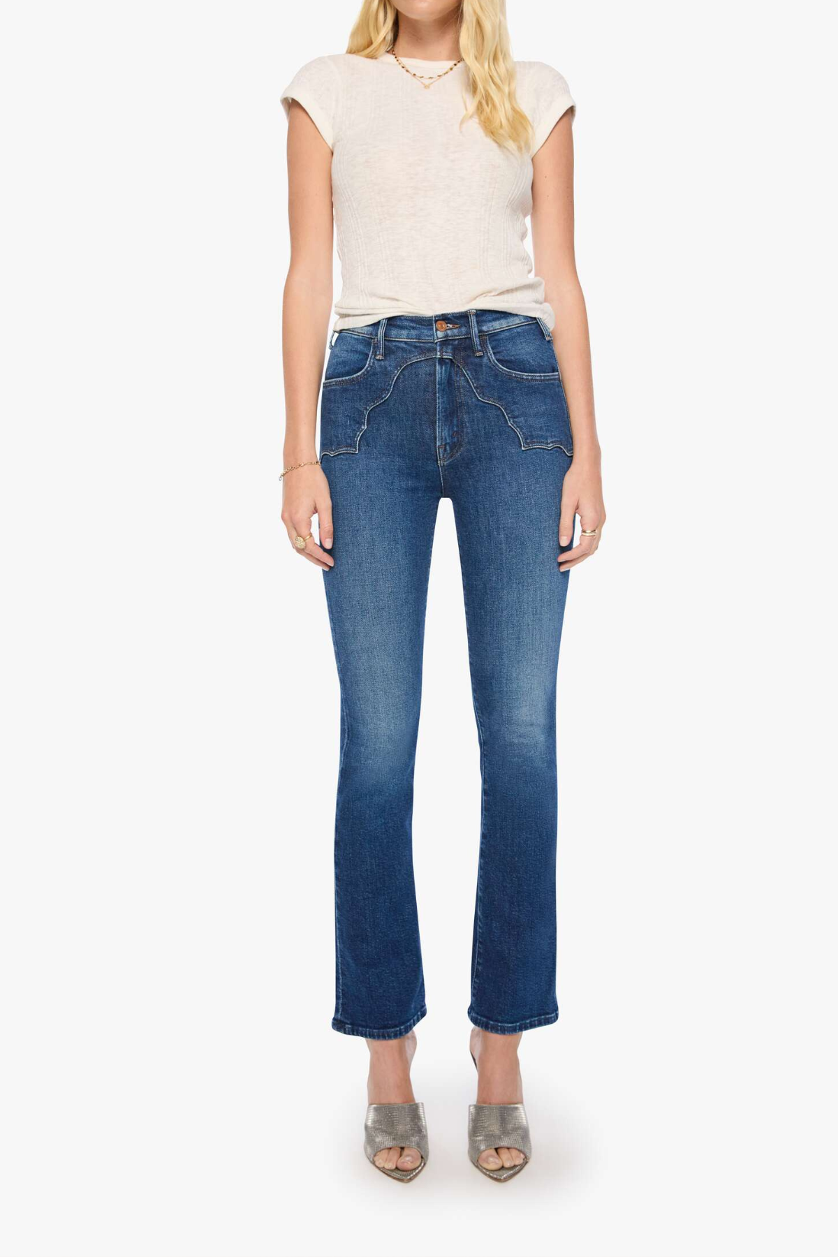 A model wearing a pair of medium-wash blue cropped flare jeans, paired with a cream-colored short-sleeve top and metallic silver high-heeled sandals, set against a plain light gray background.