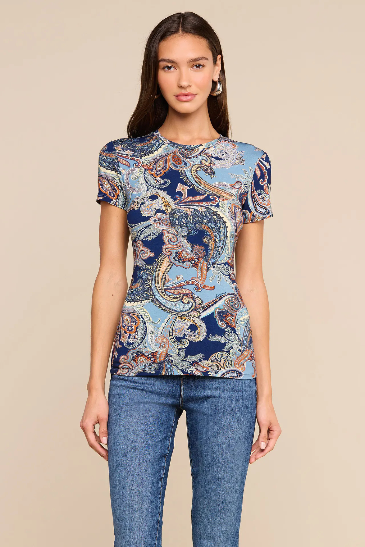 A woman modeling a short-sleeve crewneck t-shirt with a blue and beige paisley pattern, paired with blue jeans, against a solid beige background.