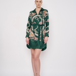 A woman modeling a dark green silk-like shirt dress with a detailed multicolor botanical and animal print, featuring a collar, button-up front, and matching tie belt, set against a plain white studio background.
