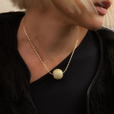 A close-up lifestyle shot of a model wearing a gold chain necklace with a textured spherical pendant, styled with a black top and black fringed jacket.