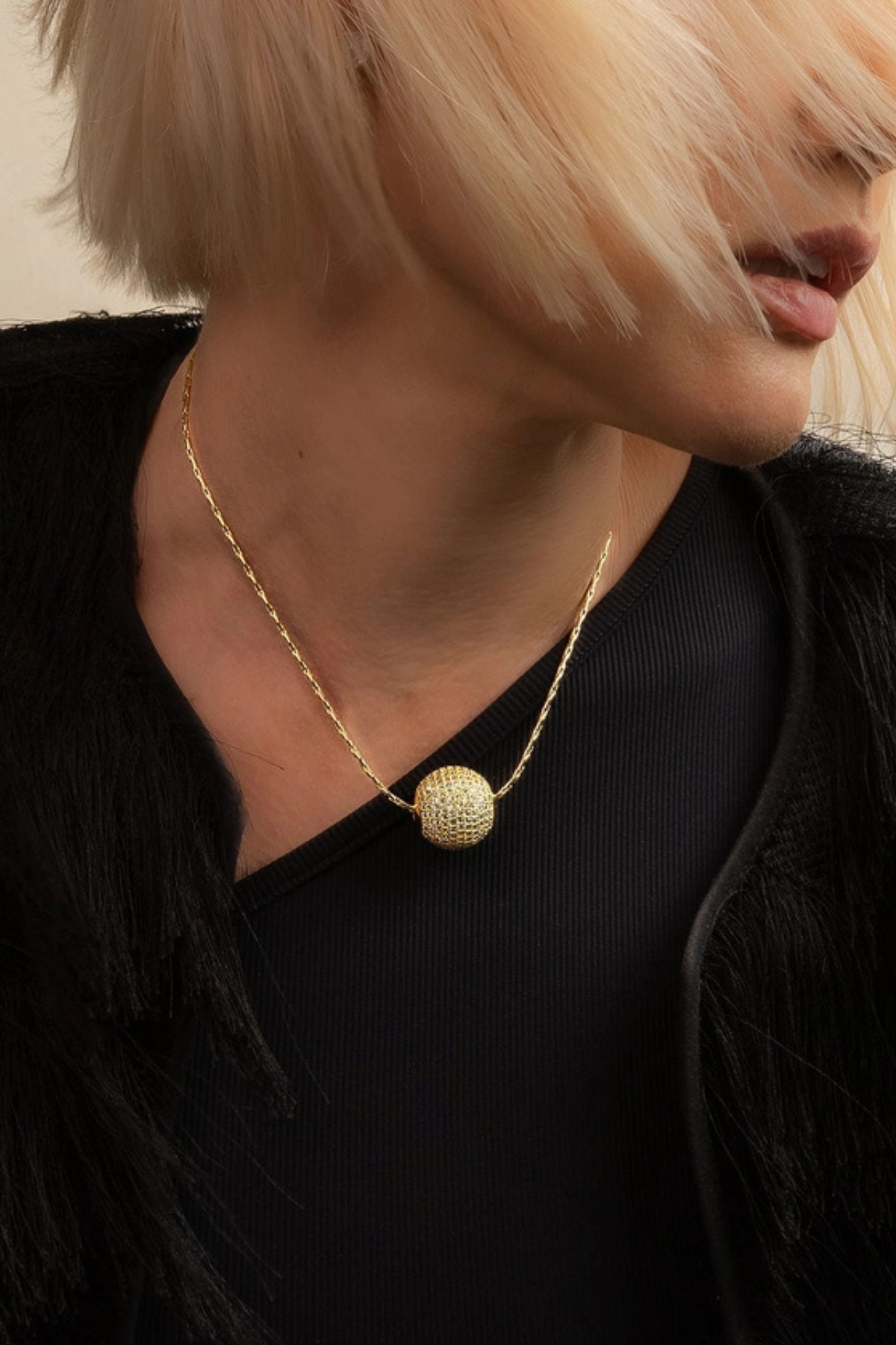A close-up lifestyle shot of a model wearing a gold chain necklace with a textured spherical pendant, styled with a black top and black fringed jacket.