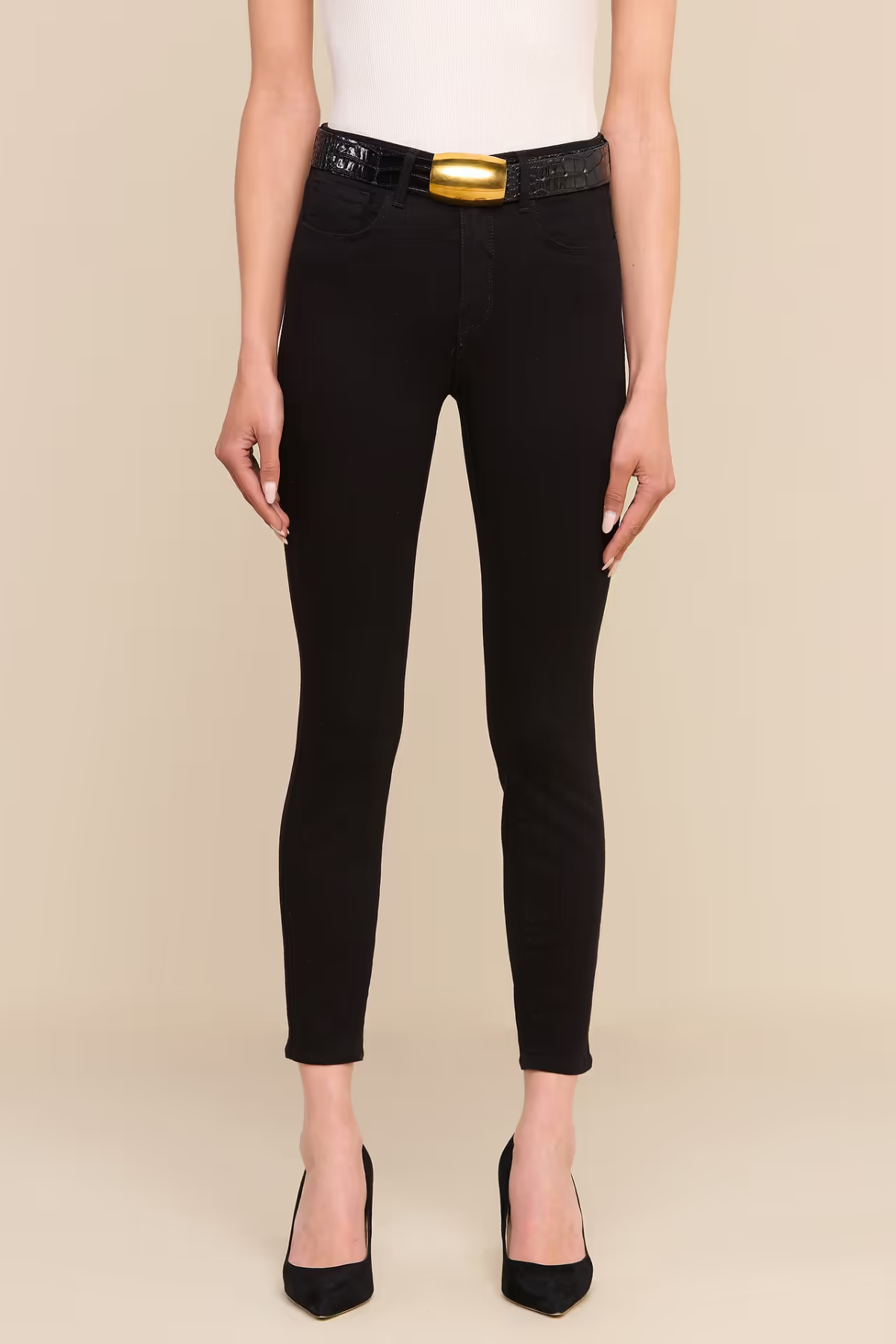 A model wearing black skinny jeans with a black belt featuring a prominent gold buckle, styled with a white top and black heels against a beige background.