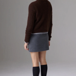 A woman shown from the back wearing a dark brown knit sweater, a gray pleated mini skirt, black knee-high socks, and black pointed-toe heels against a plain gray studio background.