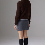 A woman shown from the back wearing a dark brown knit sweater, a gray pleated mini skirt, black knee-high socks, and black pointed-toe heels against a plain gray studio background.