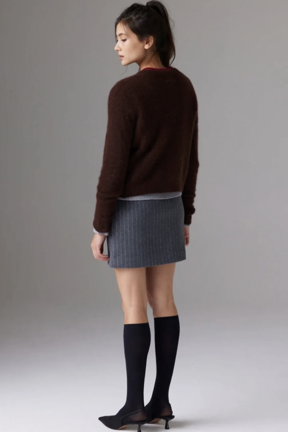 A woman shown from the back wearing a dark brown knit sweater, a gray pleated mini skirt, black knee-high socks, and black pointed-toe heels against a plain gray studio background.