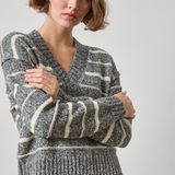A woman wearing a gray and cream striped knit sweater with a V-neck, paired with a black satin skirt, photographed against a neutral light gray background.