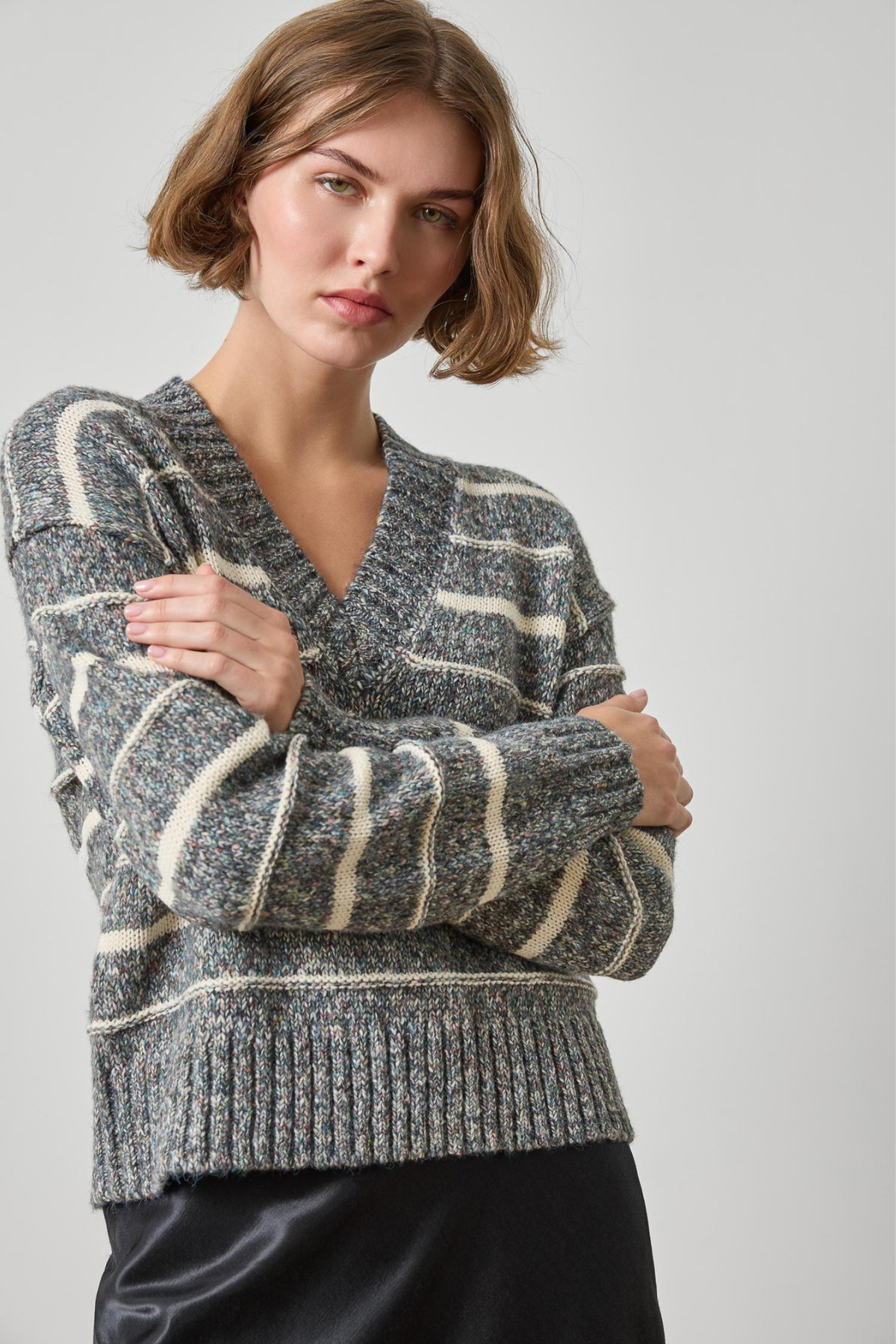 A woman wearing a gray and cream striped knit sweater with a V-neck, paired with a black satin skirt, photographed against a neutral light gray background.