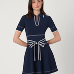 A woman modeling a short-sleeve navy blue dress with white contrast piping and a matching waist tie, photographed against a plain light gray studio background.