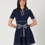 A woman modeling a short-sleeve navy blue dress with white contrast piping and a matching waist tie, photographed against a plain light gray studio background.