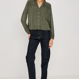 A woman modeling a long-sleeve olive green blouse with decorative pleated detailing on the shoulders and a button-down front, paired with dark blue jeans and black heeled sandals against a plain white studio background.