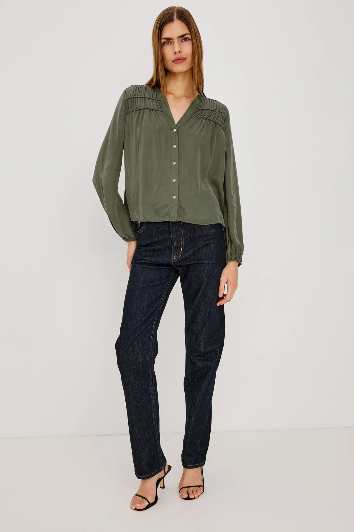A woman modeling a long-sleeve olive green blouse with decorative pleated detailing on the shoulders and a button-down front, paired with dark blue jeans and black heeled sandals against a plain white studio background.