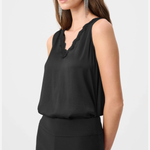 A woman modeling a black sleeveless top with a scalloped V-neckline, paired with black pants, against a light gray studio background.