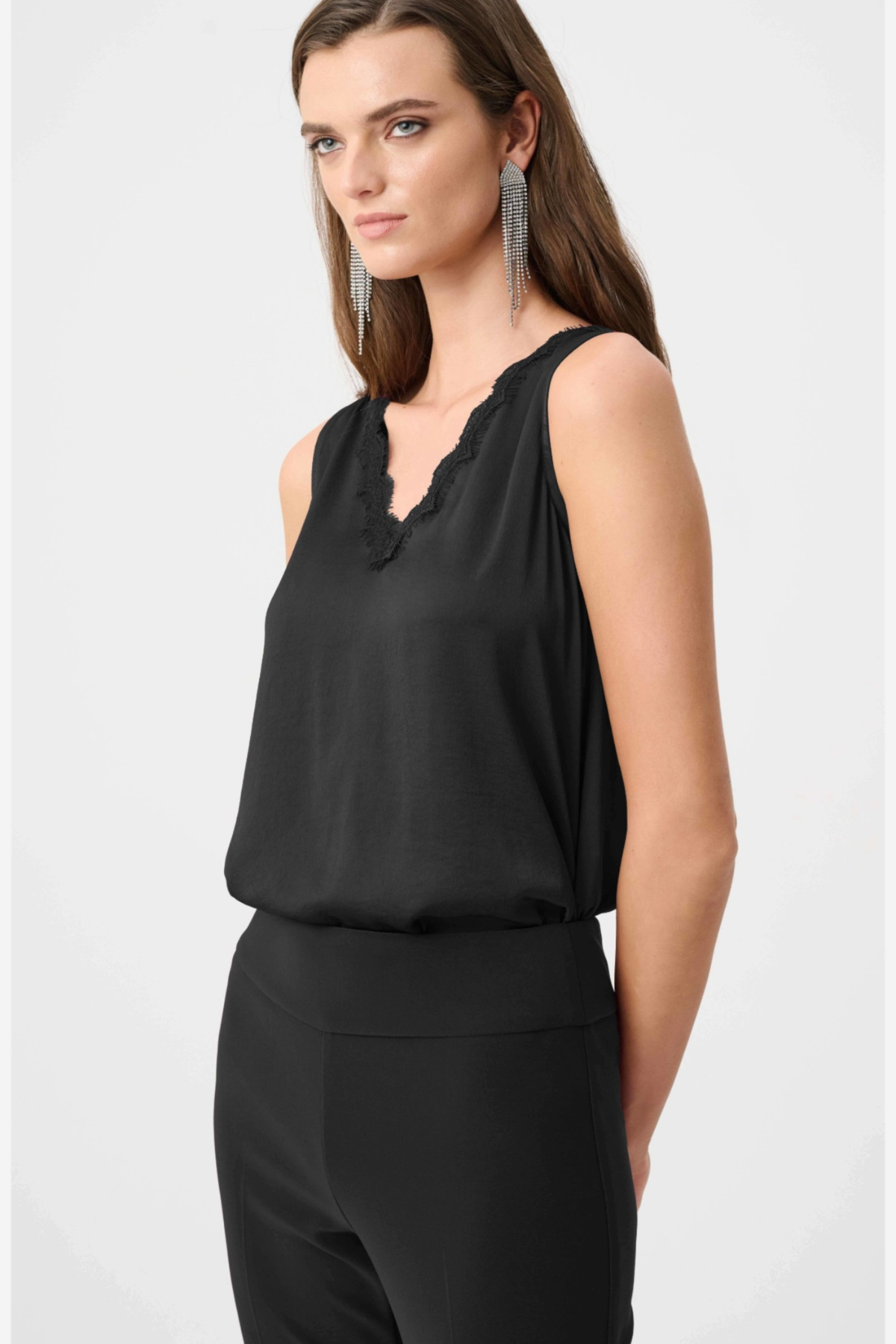 A woman modeling a black sleeveless top with a scalloped V-neckline, paired with black pants, against a light gray studio background.