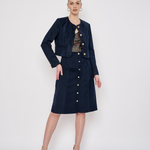 A model stands against a light gray studio background wearing a dark navy blue corduroy two-piece set consisting of a cropped jacket and a matching knee-length skirt, both detailed with gold-tone buttons. She is also wearing a patterned top and nude high-heeled sandals.