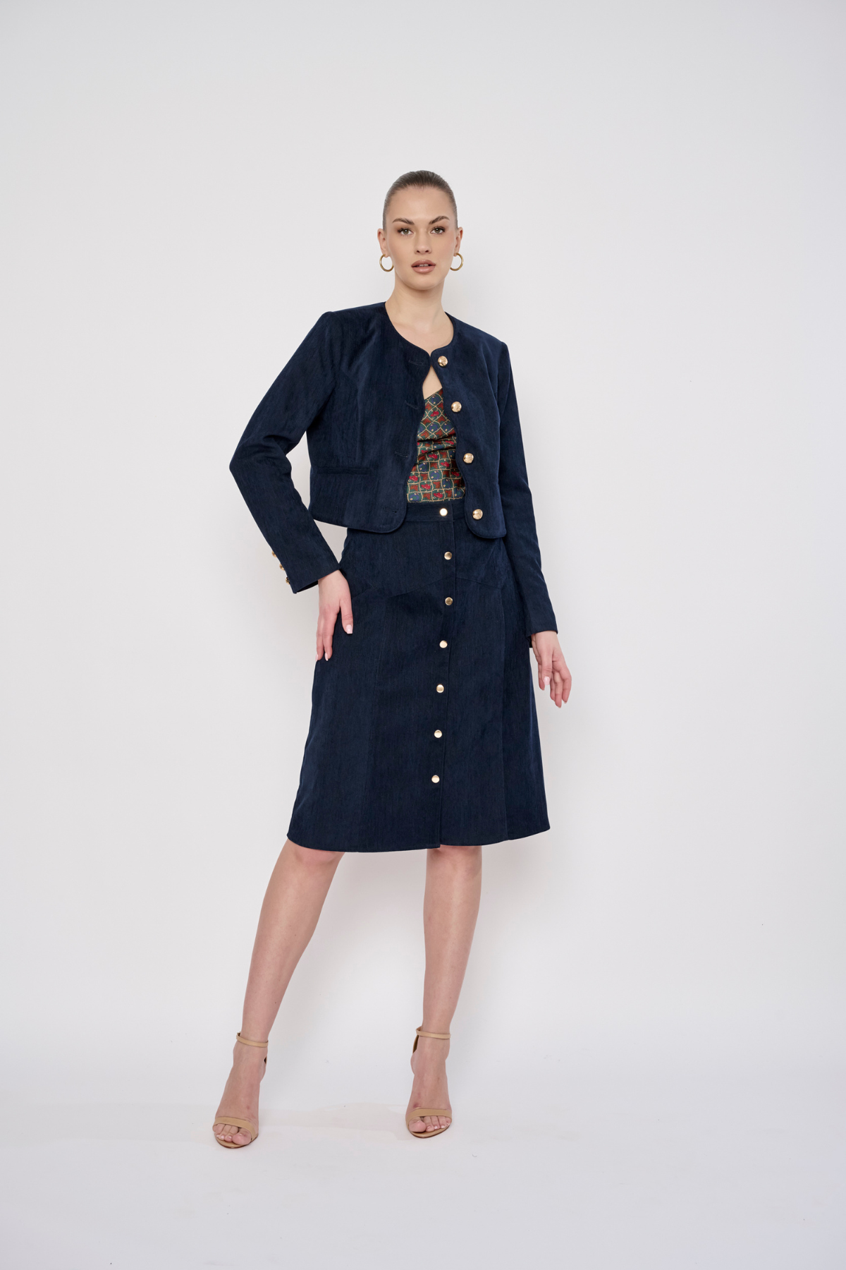 A model stands against a light gray studio background wearing a dark navy blue corduroy two-piece set consisting of a cropped jacket and a matching knee-length skirt, both detailed with gold-tone buttons. She is also wearing a patterned top and nude high-heeled sandals.