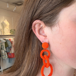 Model wearing bright orange linked drop earrings featuring three open circular shapes in a bold statement design.