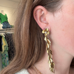 A close-up of a woman's ear wearing a long, gold leaf-shaped dangle earring with a small flower detail at the top.