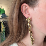 A close-up of a woman's ear wearing a long, gold leaf-shaped dangle earring with a small flower detail at the top.