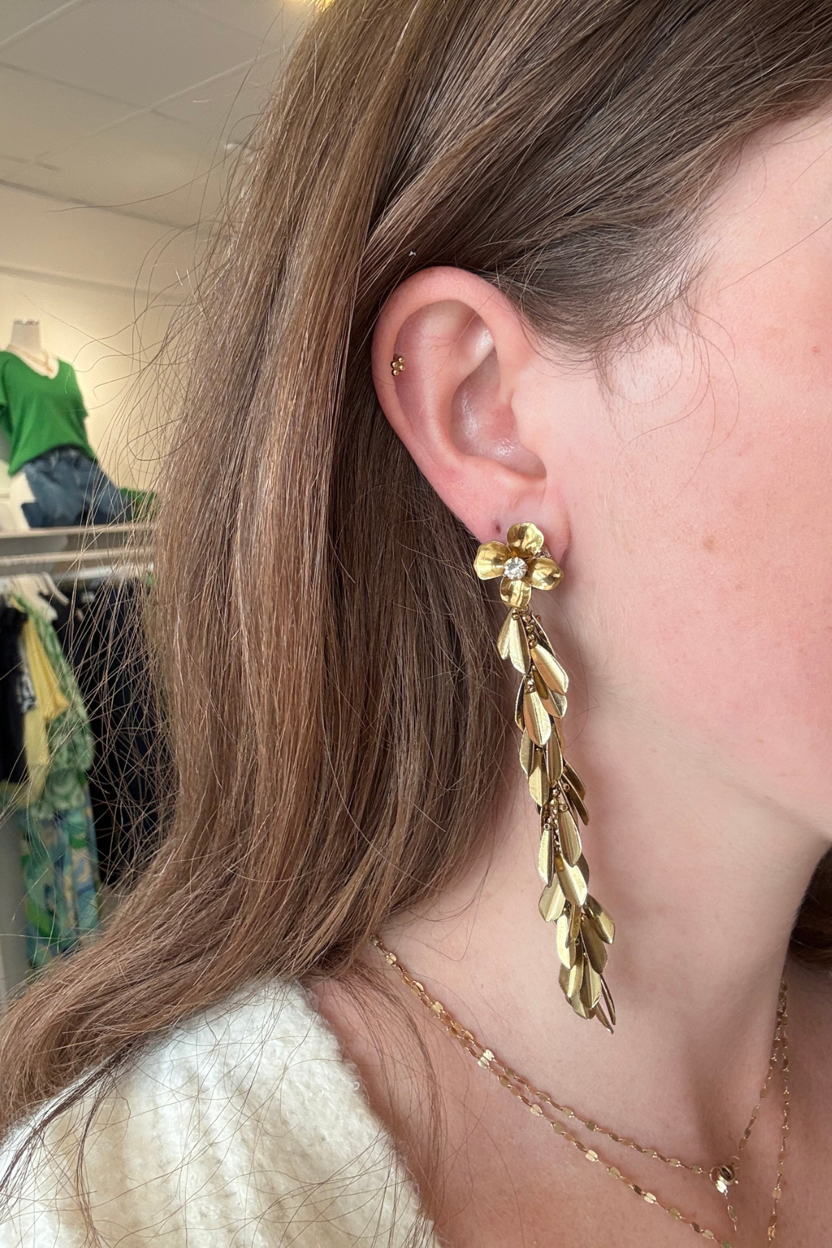 A close-up of a woman's ear wearing a long, gold leaf-shaped dangle earring with a small flower detail at the top.