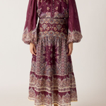 A woman wearing a long-sleeved bohemian-style midi dress in deep purple with intricate beige and white paisley patterns, paired with tan suede boots, against a plain light beige background.