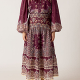 A woman wearing a long-sleeved bohemian-style midi dress in deep purple with intricate beige and white paisley patterns, paired with tan suede boots, against a plain light beige background.