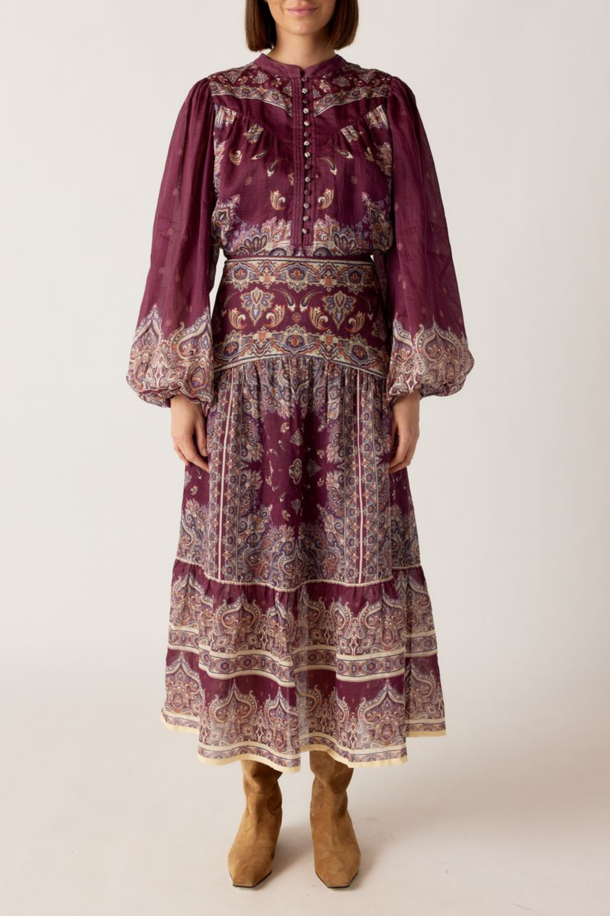 A woman wearing a long-sleeved bohemian-style midi dress in deep purple with intricate beige and white paisley patterns, paired with tan suede boots, against a plain light beige background.