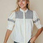 A woman wearing a cream-colored blouse with a textured gray geometric pattern across the chest and sleeve cuffs, paired with blue jeans, set against a plain beige background.