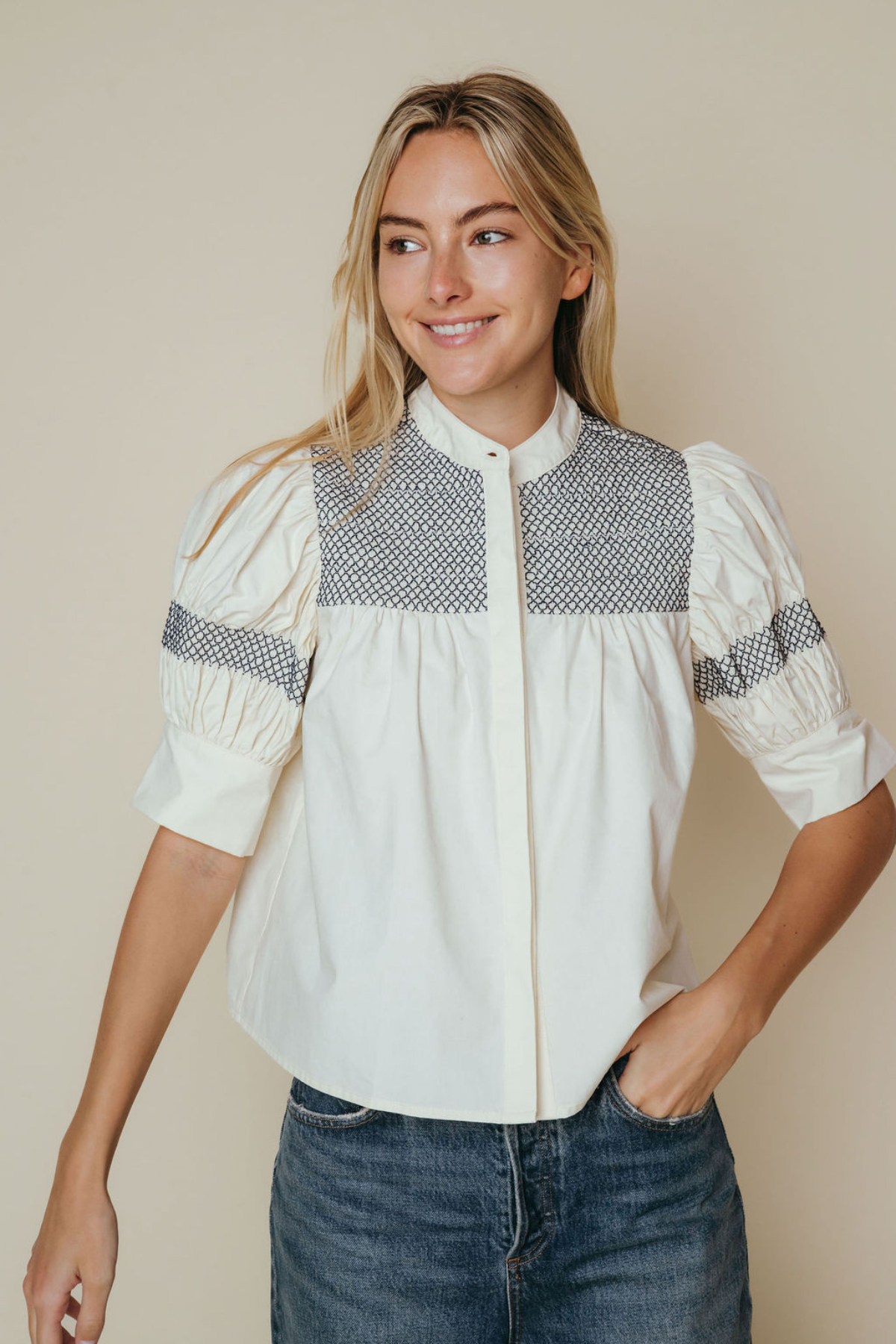 A woman wearing a cream-colored blouse with a textured gray geometric pattern across the chest and sleeve cuffs, paired with blue jeans, set against a plain beige background.