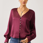 A woman modeling a long-sleeve satin blouse in deep burgundy with a button-down front and gathered neckline, paired with blue jeans against a light gray studio background.