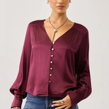 A woman modeling a long-sleeve satin blouse in deep burgundy with a button-down front and gathered neckline, paired with blue jeans against a light gray studio background.