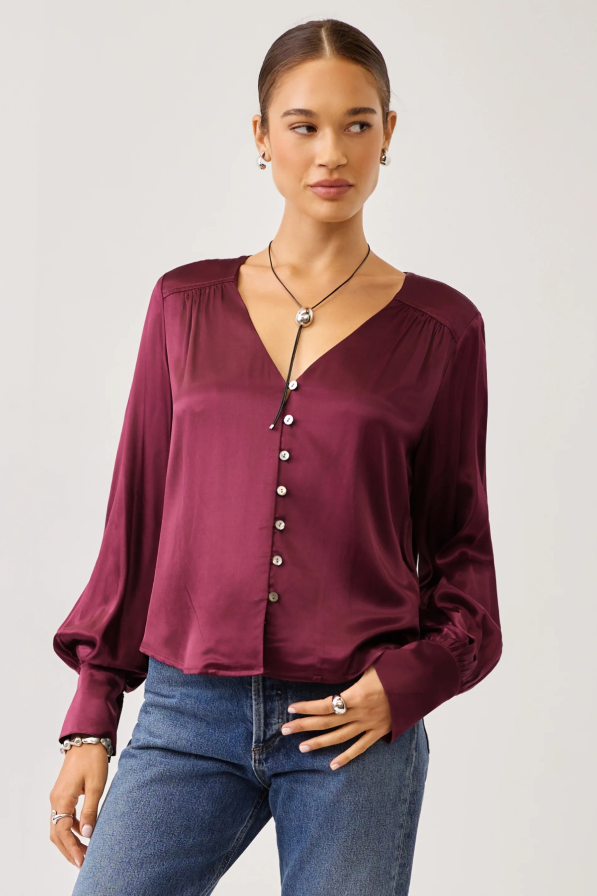 A woman modeling a long-sleeve satin blouse in deep burgundy with a button-down front and gathered neckline, paired with blue jeans against a light gray studio background.