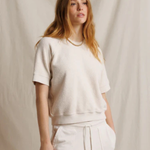 A woman modeling a cream-colored short-sleeve knit top and matching cream drawstring pants against a draped beige fabric backdrop.