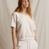 A woman modeling a cream-colored short-sleeve knit top and matching cream drawstring pants against a draped beige fabric backdrop.