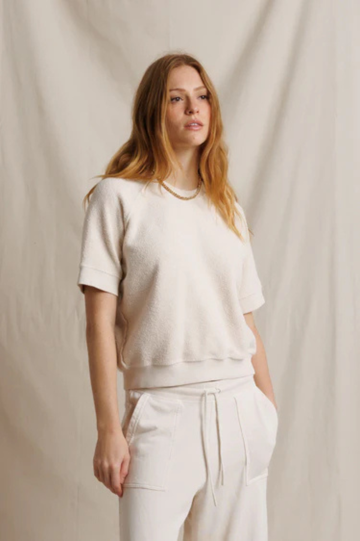 A woman modeling a cream-colored short-sleeve knit top and matching cream drawstring pants against a draped beige fabric backdrop.