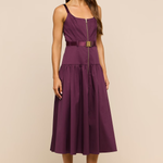 A woman modeling a sleeveless midi dress in deep plum purple with a matching waist belt and a gold-tone front zipper. She is standing against a solid beige studio background.