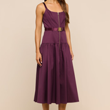 A woman modeling a sleeveless midi dress in deep plum purple with a matching waist belt and a gold-tone front zipper. She is standing against a solid beige studio background.