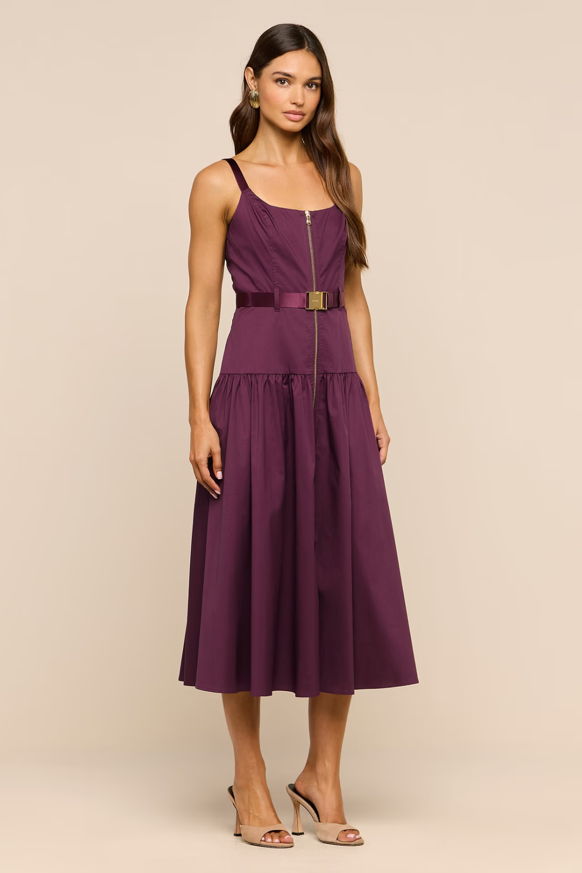 A woman modeling a sleeveless midi dress in deep plum purple with a matching waist belt and a gold-tone front zipper. She is standing against a solid beige studio background.