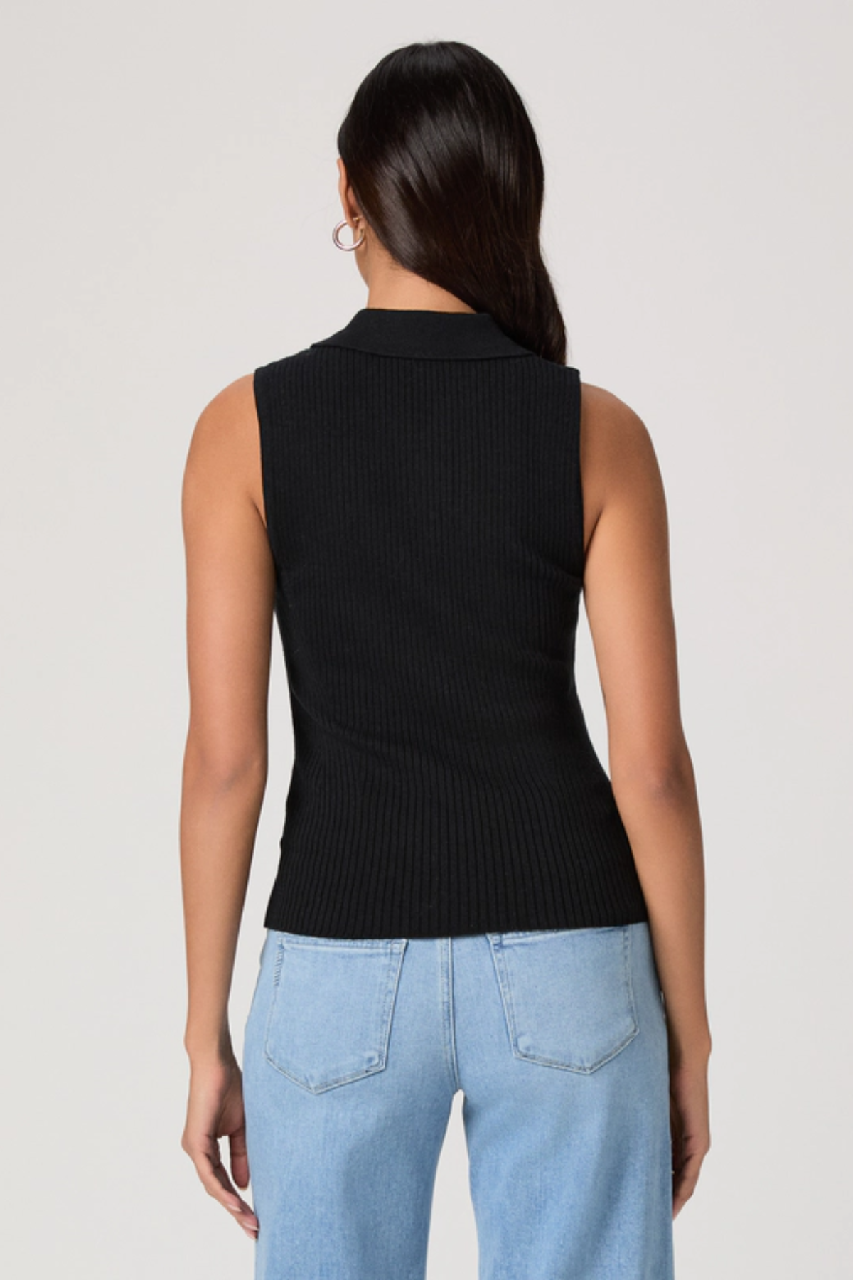 Woman wearing a black sleeveless knitted top and light blue jeans on a plain background