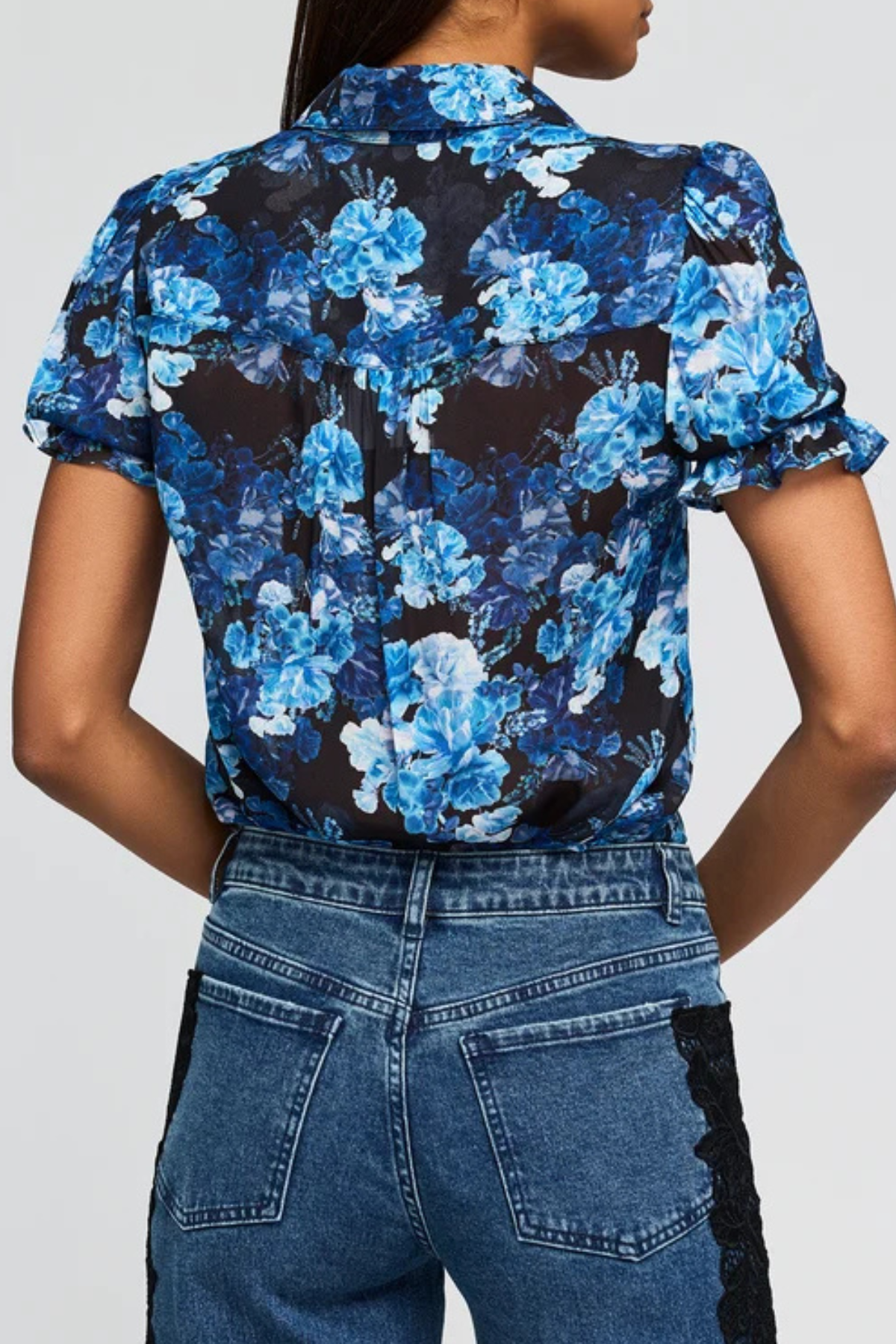 A woman shown from the back wearing a short-sleeve button-up blouse with a blue and black floral print, paired with blue jeans.