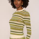 A woman with an afro hairstyle wearing a long-sleeve knit top with horizontal stripes in olive green, beige, and white, photographed against a plain light background.
