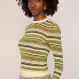 A woman with an afro hairstyle wearing a long-sleeve knit top with horizontal stripes in olive green, beige, and white, photographed against a plain light background.