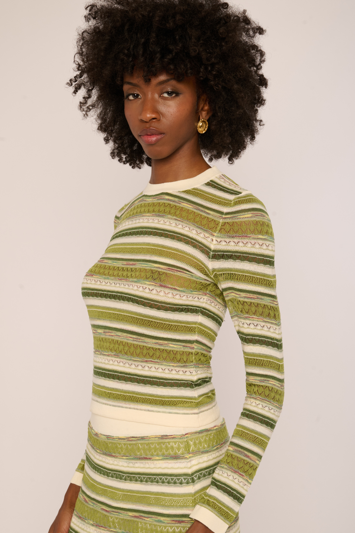 A woman with an afro hairstyle wearing a long-sleeve knit top with horizontal stripes in olive green, beige, and white, photographed against a plain light background.