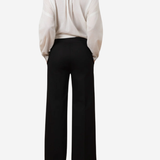 A model shown from the back wearing black wide-leg trousers and a cream-colored blouse, photographed against a solid light gray background.
