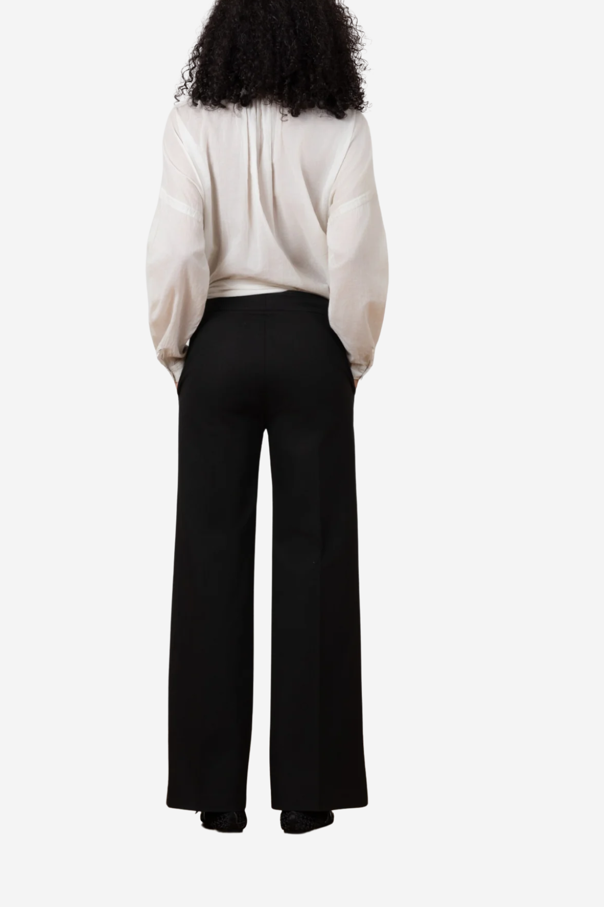 A model shown from the back wearing black wide-leg trousers and a cream-colored blouse, photographed against a solid light gray background.