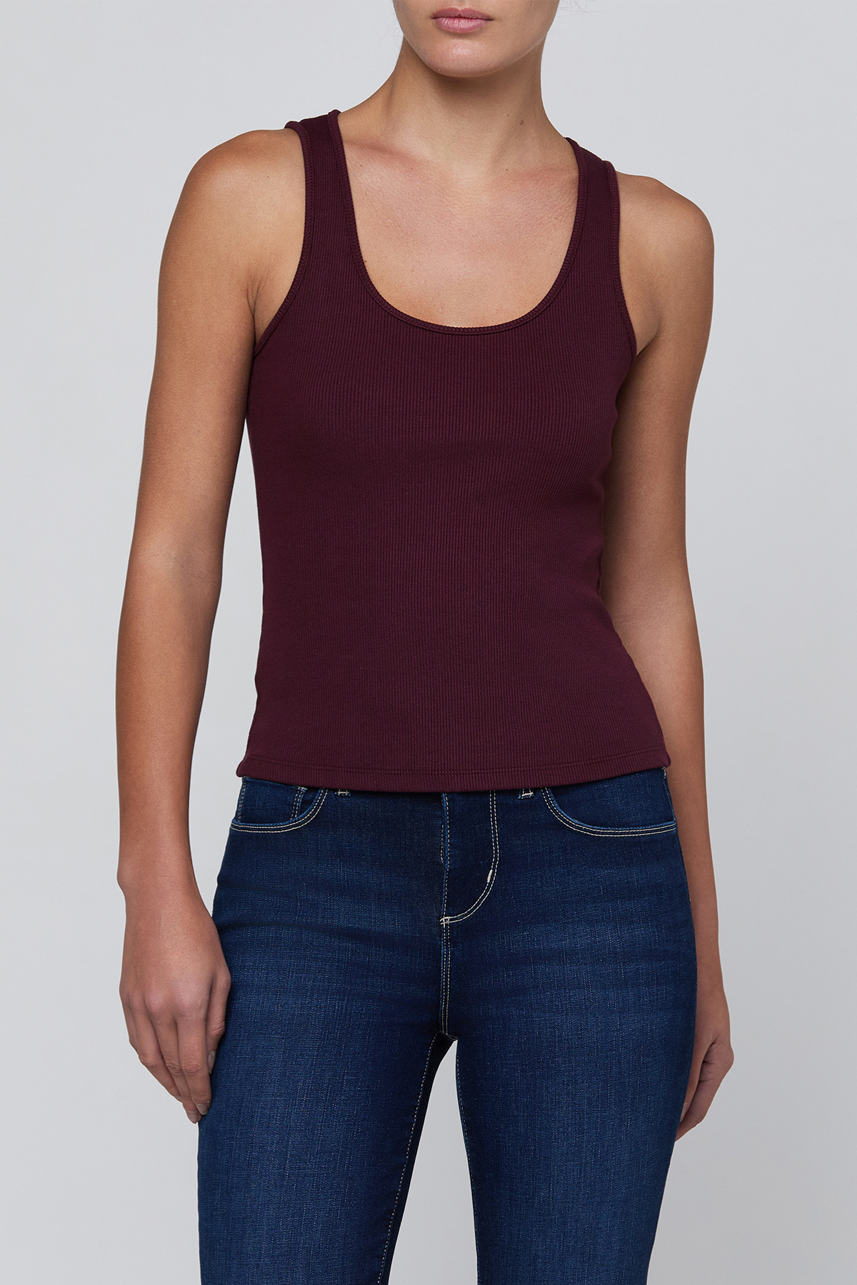 A woman wearing a sleeveless scoop-neck tank top in deep burgundy and dark blue jeans, shown from the neck down against a light gray studio background.